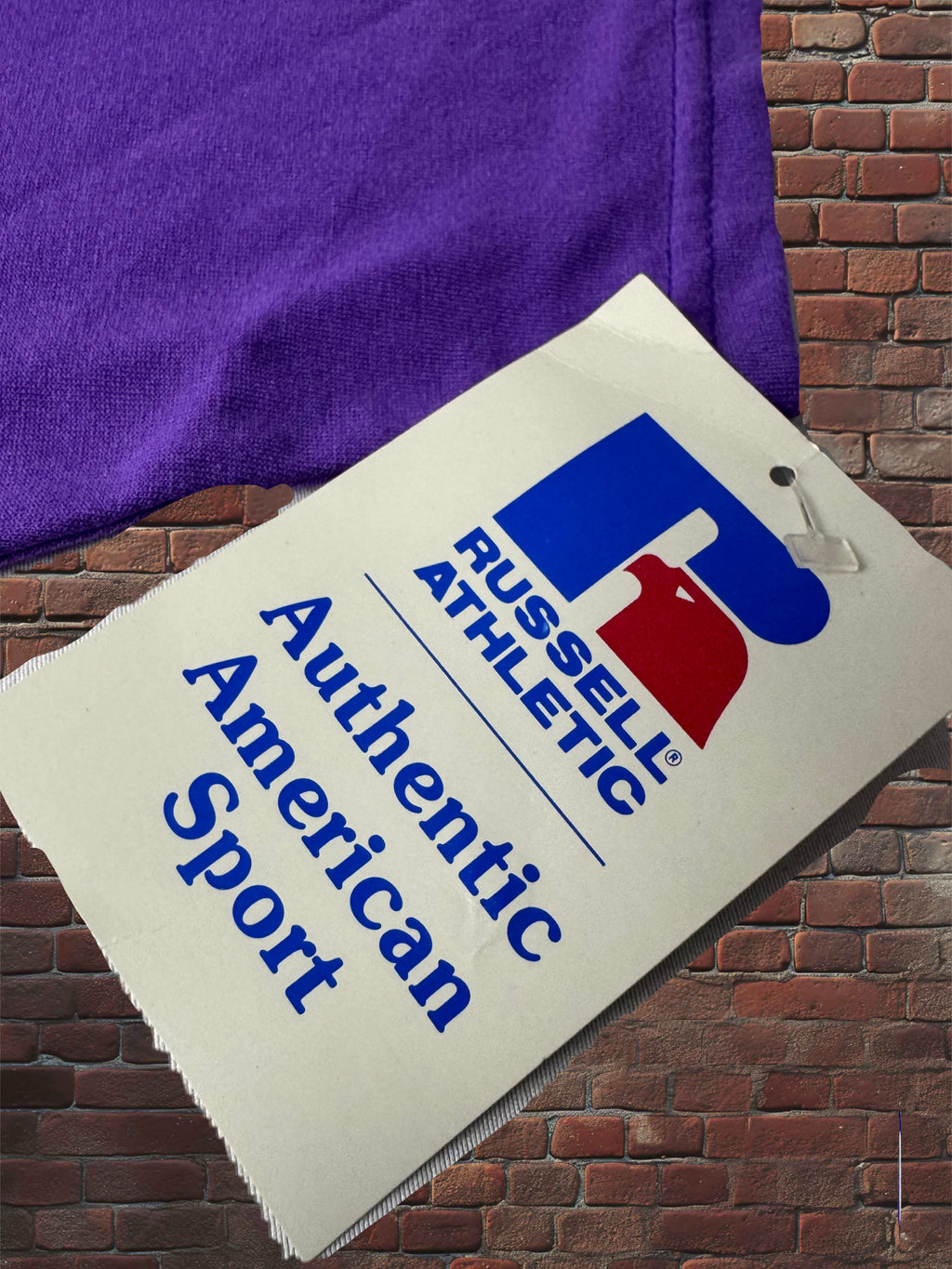 A close-up of a purple athletic fabric item, likely a sweatshirt or shirt, with a hanging paper tag that reads 'RUSSELL ATHLETIC' and 'Authentic American Sport' on a brick wall background.