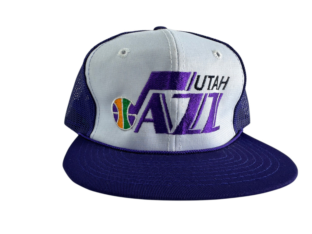 A Utah Jazz trucker hat featuring a white front panel with purple and black embroidered logo, a purple brim, and purple mesh side panels.