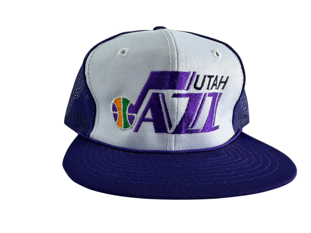 A Utah Jazz trucker hat featuring a white front panel with purple and black embroidered logo, a purple brim, and purple mesh side panels.
