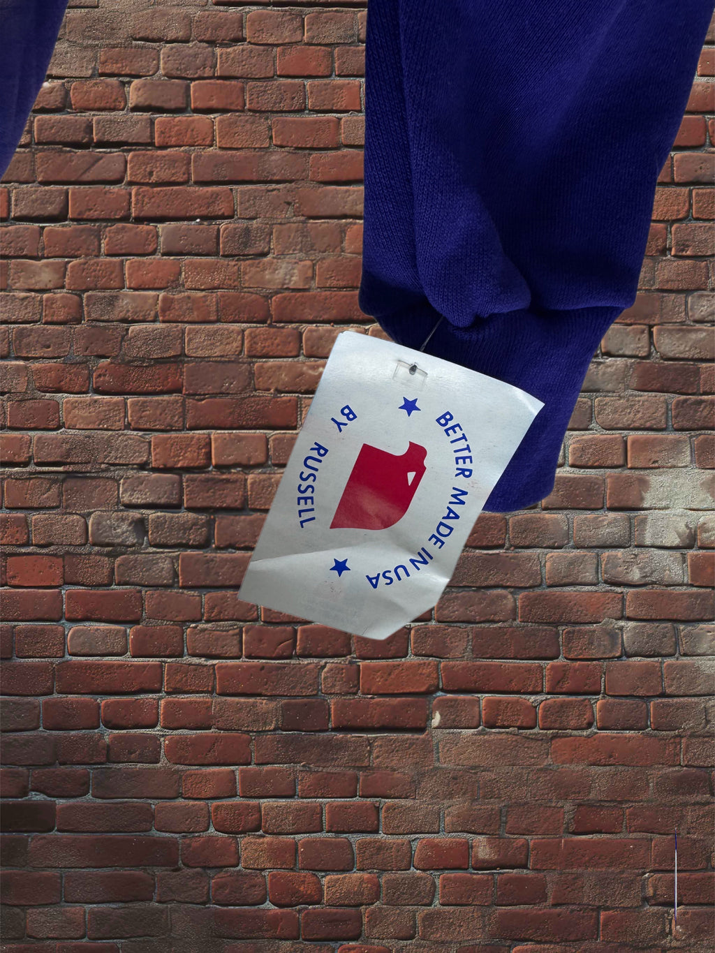 A close-up of a dark blue knit garment hanging against a red-brown brick wall, with a white paper hangtag attached. The tag reads 'BETTER MADE IN USA' and 'BY RUSSELL' with a red logo.