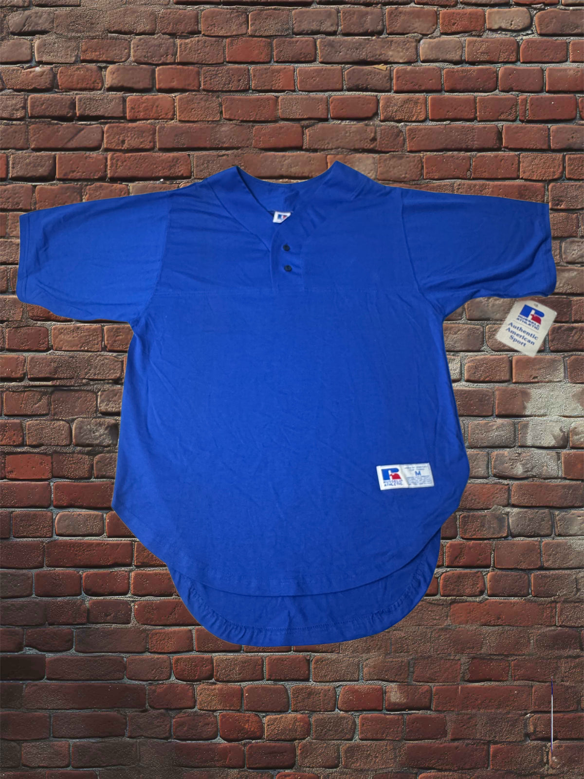 A royal blue short-sleeve henley-style baseball shirt laid flat against a red-brown brick wall background. The shirt has a three-button placket, a curved hem, and a small white brand tag near the lower right front, with a smaller hangtag attached to the right sleeve.