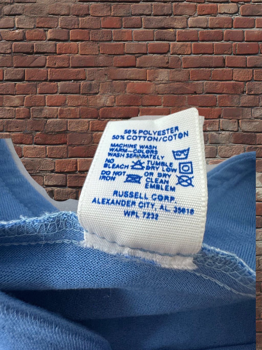 A close-up of a blue garment's interior care label sewn onto the collar, with a brick wall background.