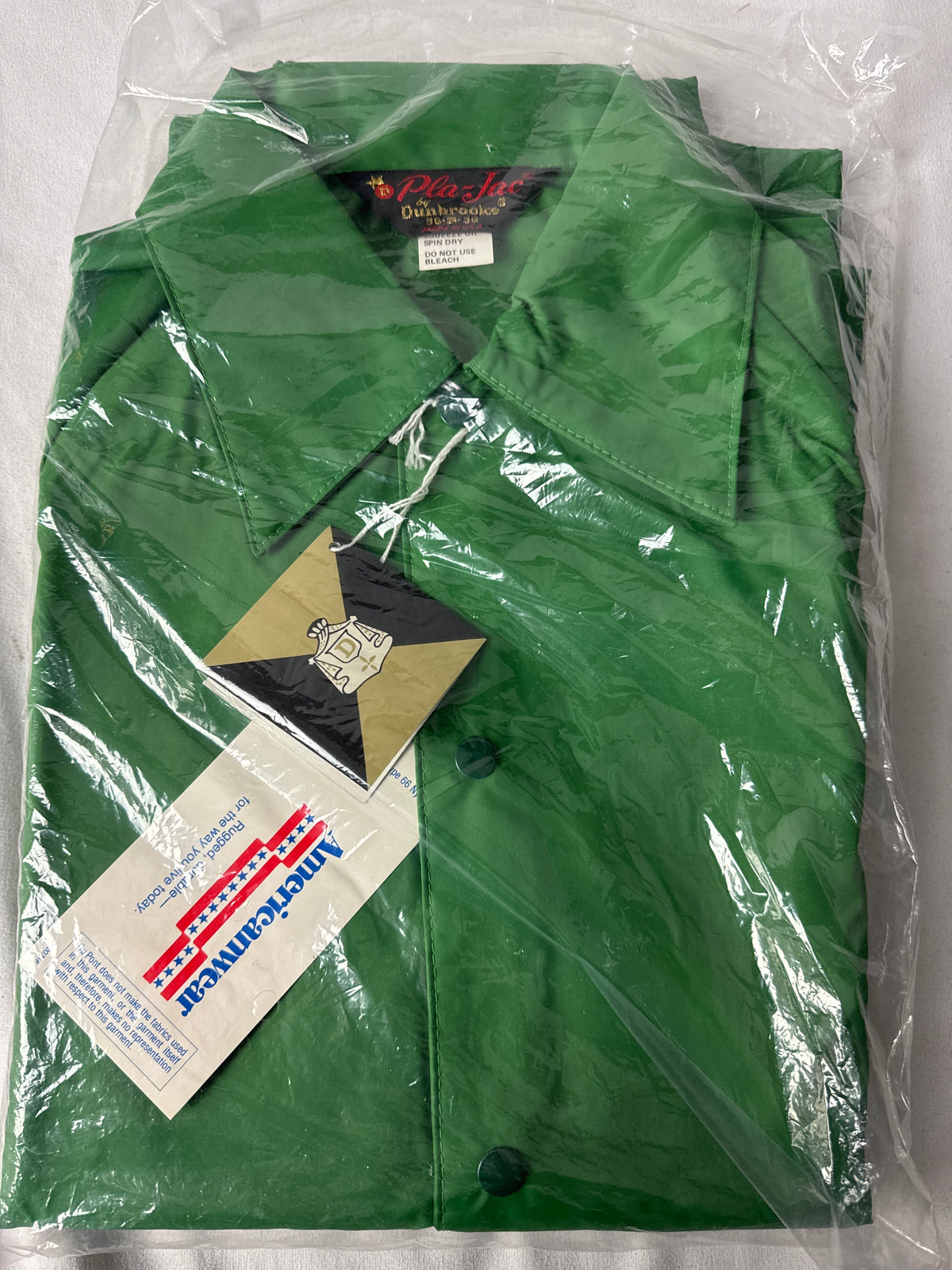A folded green nylon jacket inside a clear plastic bag, with a paper hangtag and a triangular brand tag attached. The jacket has a pointed collar and dark buttons.
