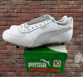 A white Puma Rocket 2 cleat shoe resting on top of its green original box, set against a brick wall background.