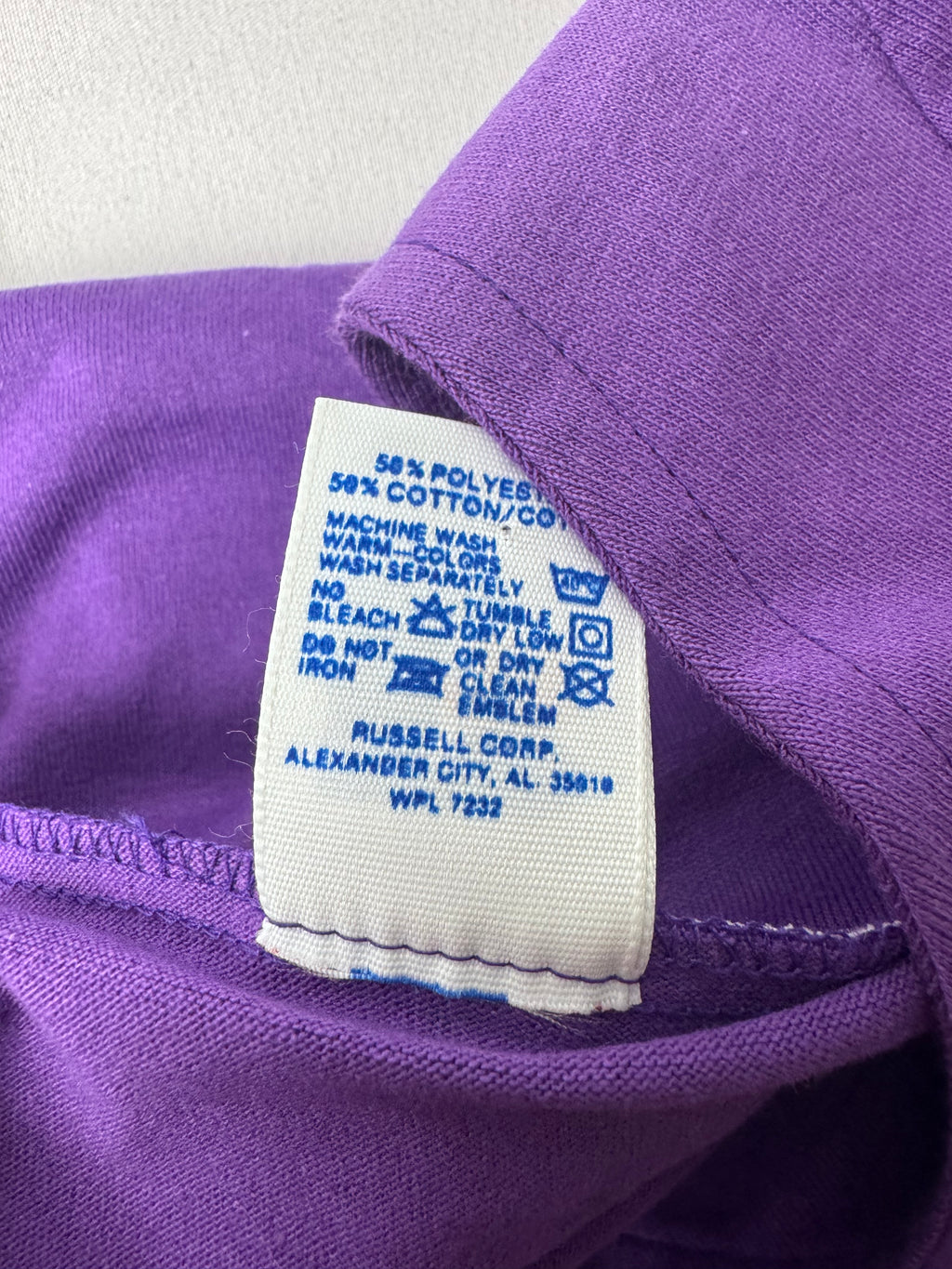 Close-up of a purple garment's collar showing a white fabric care label with blue text and washing instructions. The label reads '50% POLYESTER 50% COTTON/CPL' and 'RUSSELL CORP.