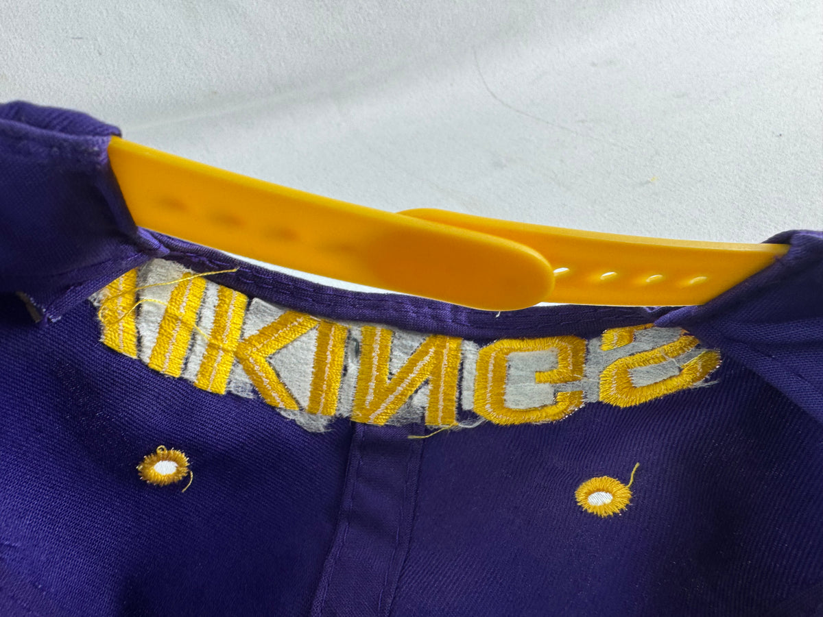 Close-up of a purple and gold baseball cap showing the embroidered Vikings' wordmark on the back, a gold plastic snap strap, and the purple fabric crown.