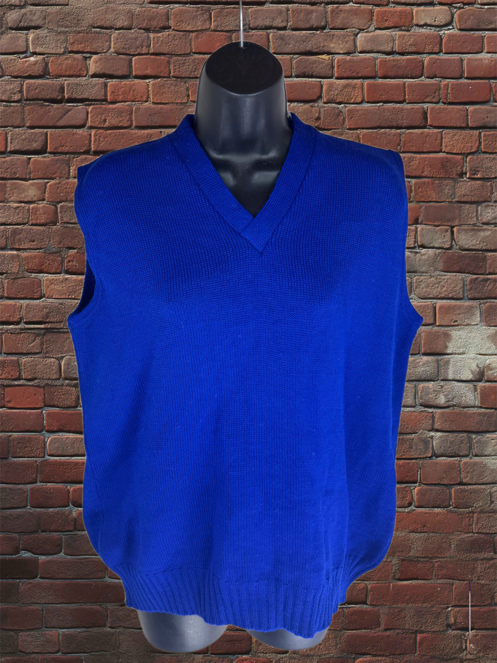 A bright royal blue ribbed knit sweater vest displayed on a black mannequin torso, set against a red-brown brick wall background.
