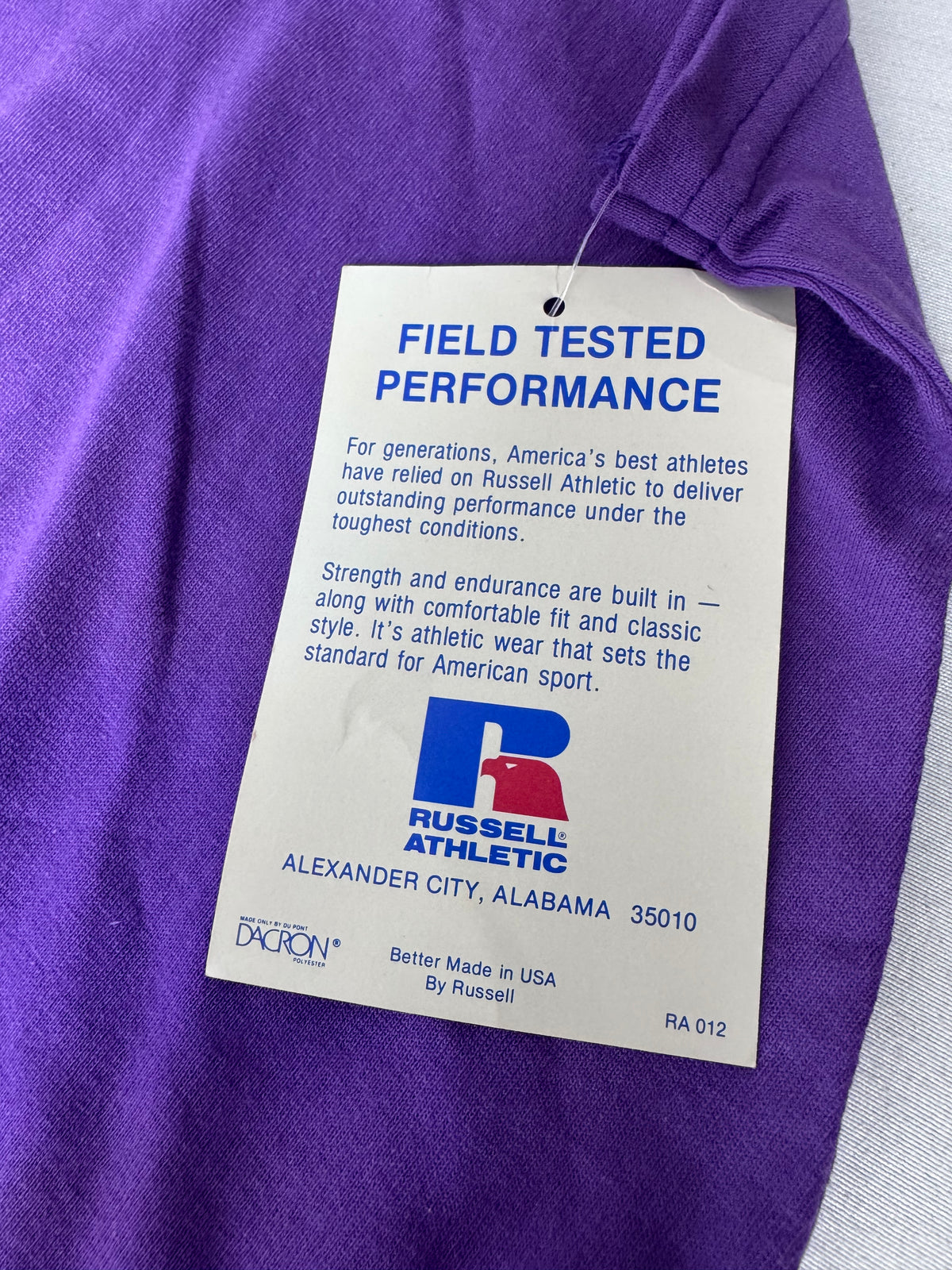 A close-up of a purple athletic shirt with a white hangtag reading 'FIELD TESTED PERFORMANCE' and showing the Russell Athletic logo and address.