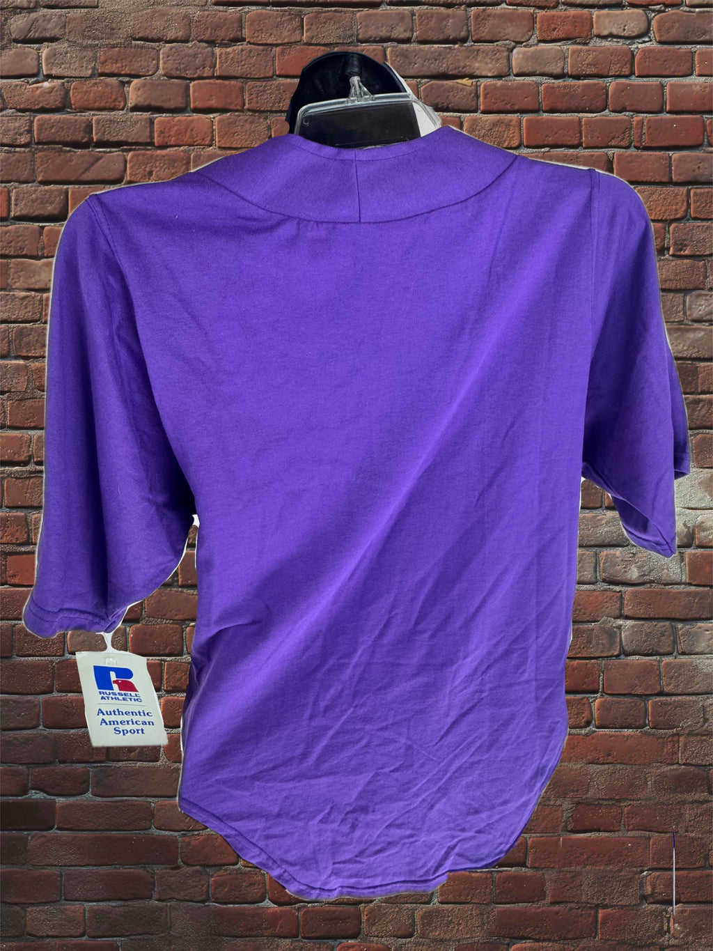 A purple short-sleeve t-shirt shown from the back on a hanger against a red brick wall background, with a white authenticity tag hanging from the sleeve.