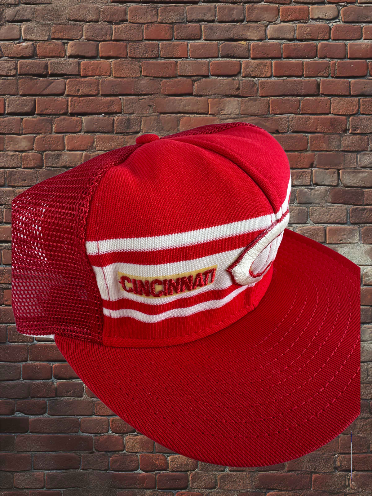 A red trucker-style baseball cap with white stripes and a white embroidered patch reading CINCINNATI, shown against a brick wall background.