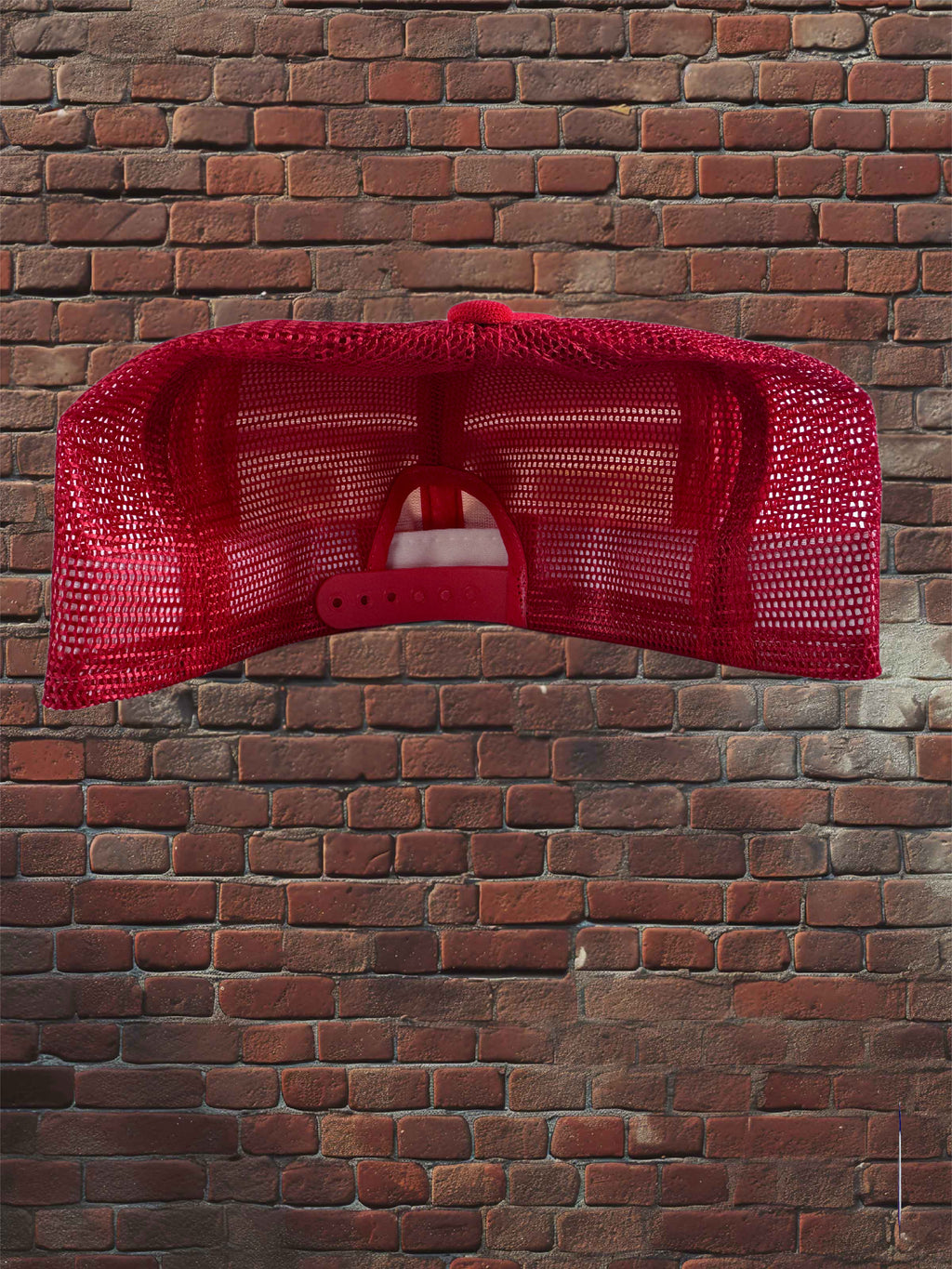 A bright red trucker-style cap with a mesh back, shown from the back against a red brick wall background.