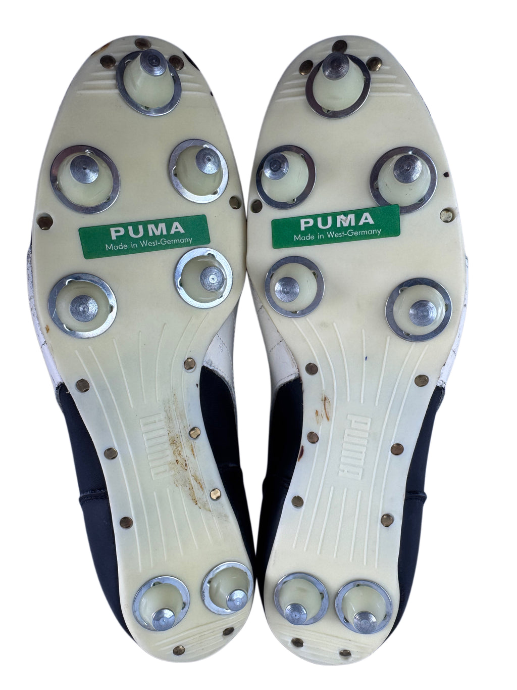 The soles of a pair of Puma athletic cleats, showing the off-white plastic sole with multiple plastic cleats with metal tips  and green Puma logos.