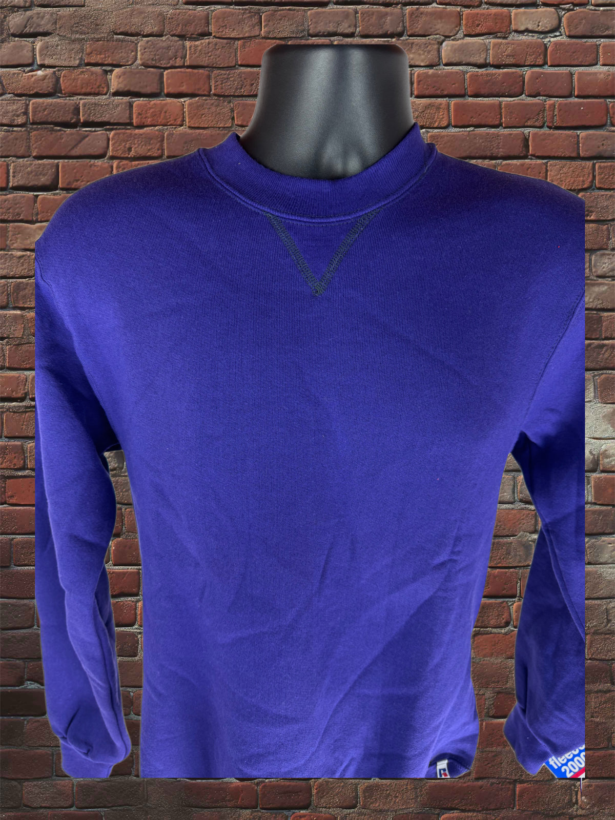 A royal blue long-sleeve crewneck shirt displayed on a black mannequin torso against a red brick wall background.