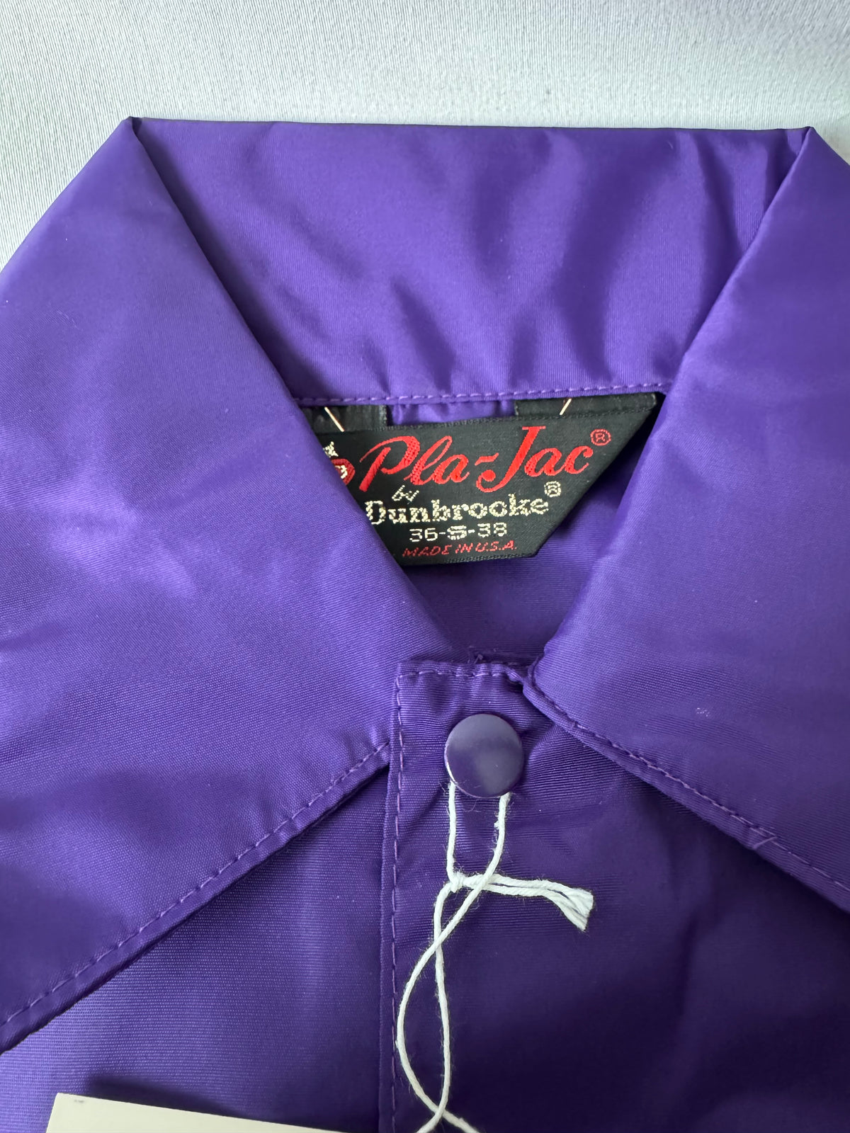 Close-up of a purple collared work jacket or with a visible inner neck label reading 'Pla-Jac by Dunbrooke' and a white hanging tag tied to the collar.
