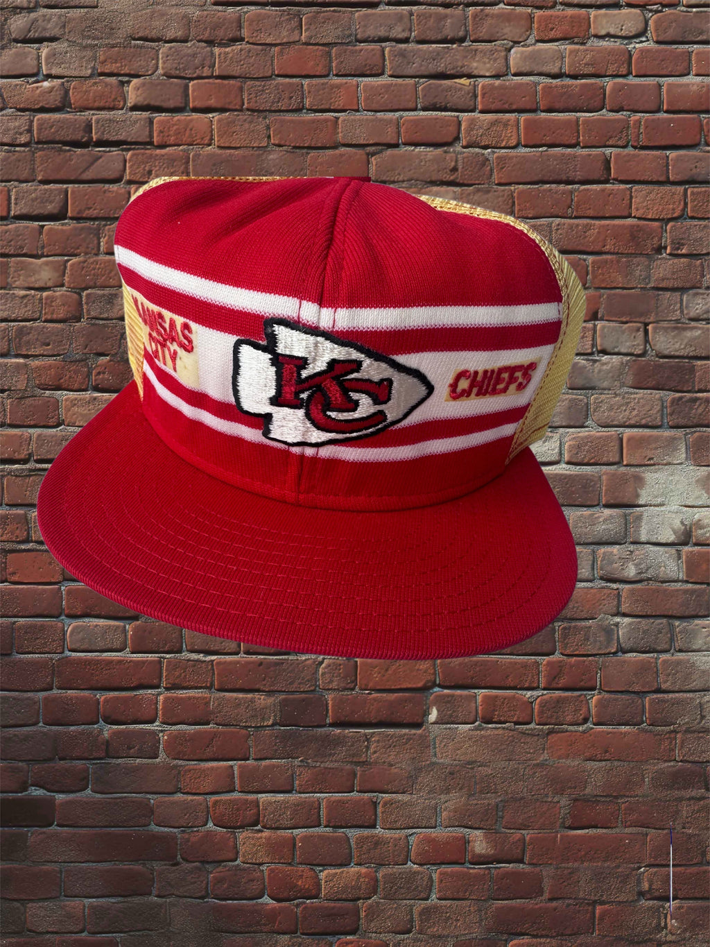 A red Kansas City Chiefs trucker hat with a red flat brim, white and red horizontal stripes on the front, and a gold mesh back, set against a red brick wall background.