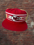 A red Kansas City Chiefs trucker hat with a red flat brim, white and red horizontal stripes on the front, and a gold mesh back, set against a red brick wall background.
