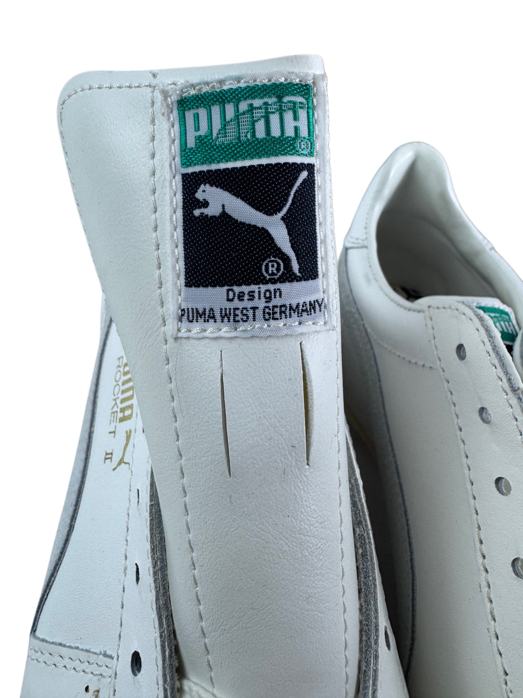 Close-up of a white Puma shoe showing the heel tab with the green and black Puma West Germany logo patch, alongside part of the side panel and eyelets.