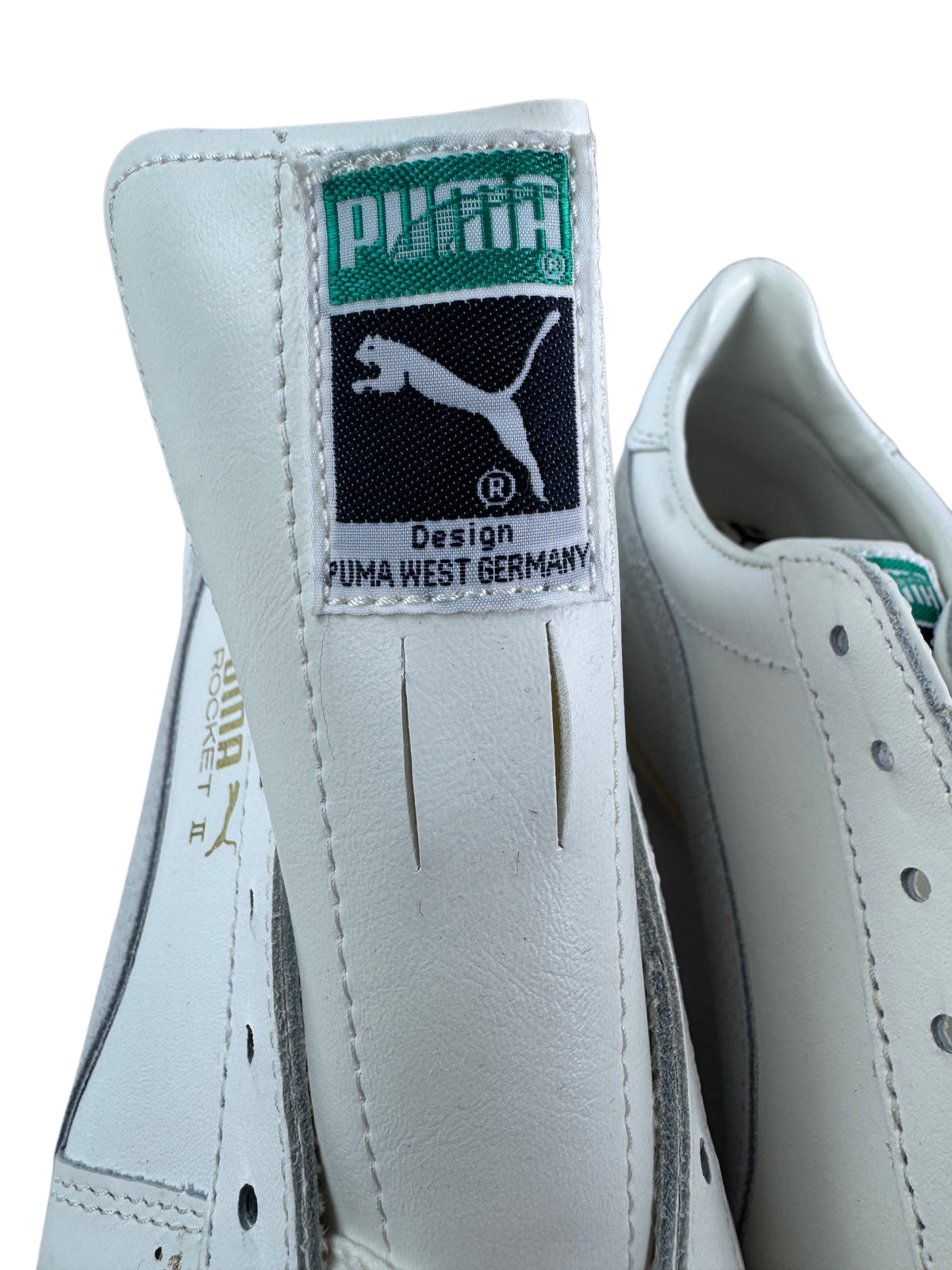 Close-up of a white Puma shoe showing the heel tab with the green and black Puma West Germany logo patch, alongside part of the side panel and eyelets.
