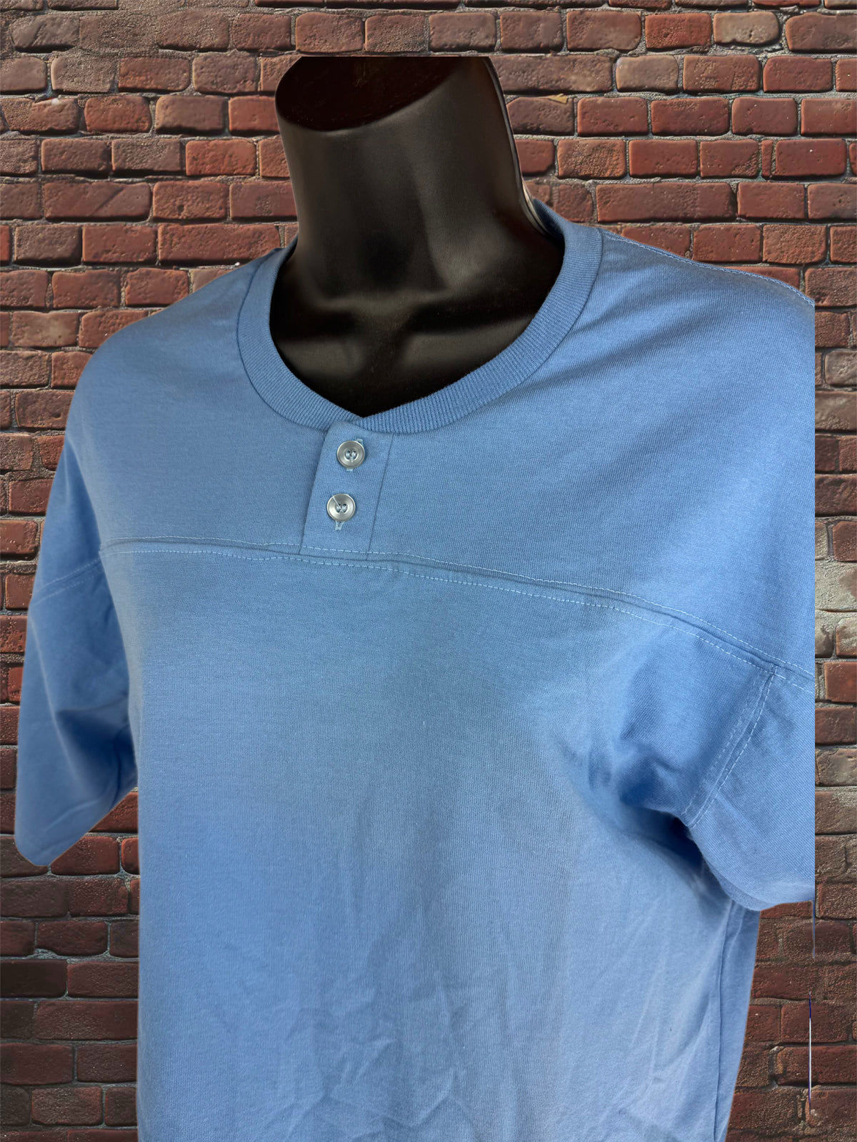 A light blue short-sleeve henley t-shirt displayed on a black mannequin torso, set against a red-brown brick wall background.