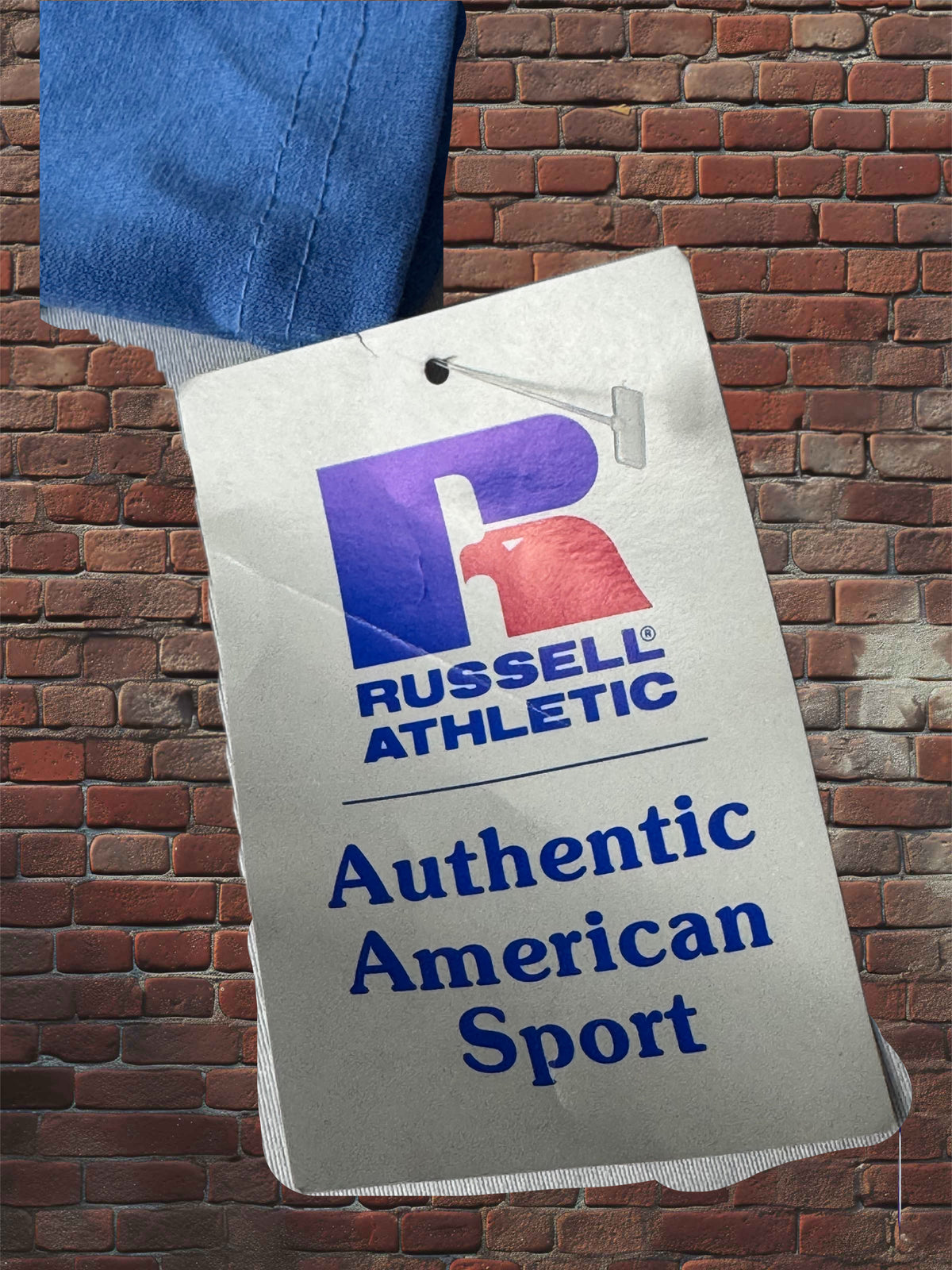 A close-up of a Russell Athletic hangtag reading 'Authentic American Sport' against a red brick wall background, with a blue fabric corner visible at the top left.