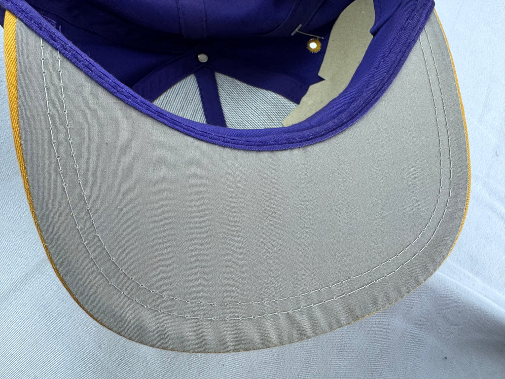 Close-up of a baseball cap showing the curved brim, light gray fabric, white stitching, purple inner lining, and a thin yellow edge trim.