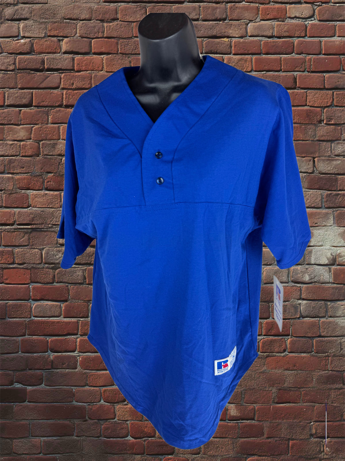 A royal blue short-sleeve baseball-style polo shirt displayed on a black mannequin torso against a red-brown brick wall background.