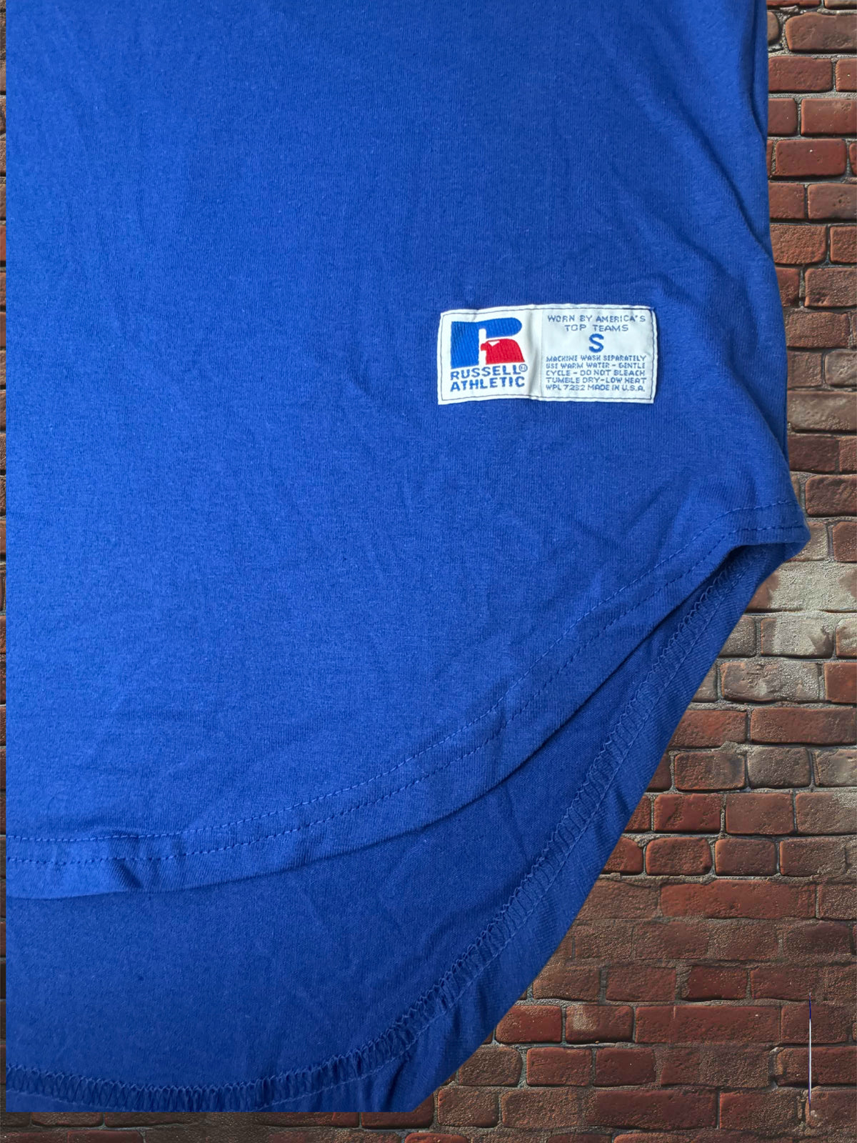 A close-up of a royal blue athletic t-shirt laid flat against a red brick wall background. The shirt features a white sewn-in label on the lower left front with the 'Russell Athletic' logo and size 'S' printed on it.