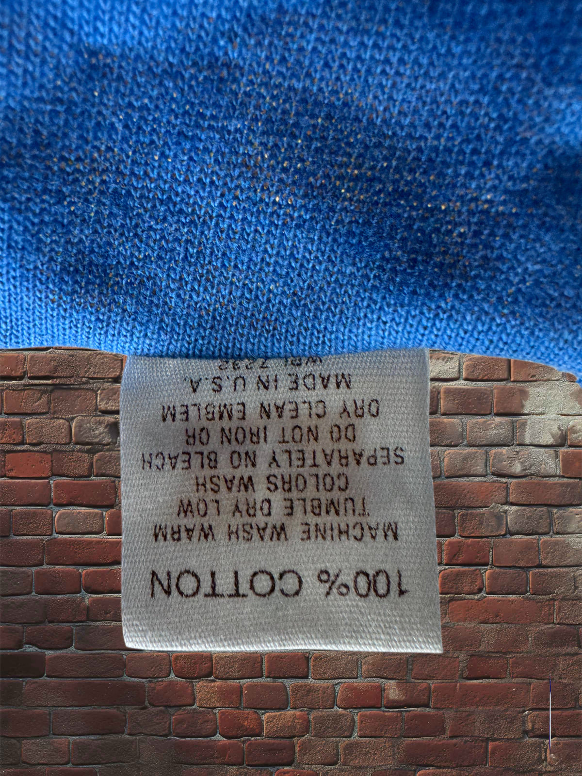 A close-up of a white cotton care label sewn onto a blue garment, set against a red-brown brick wall background. The label reads '100% COTTON' and other care instructions.
