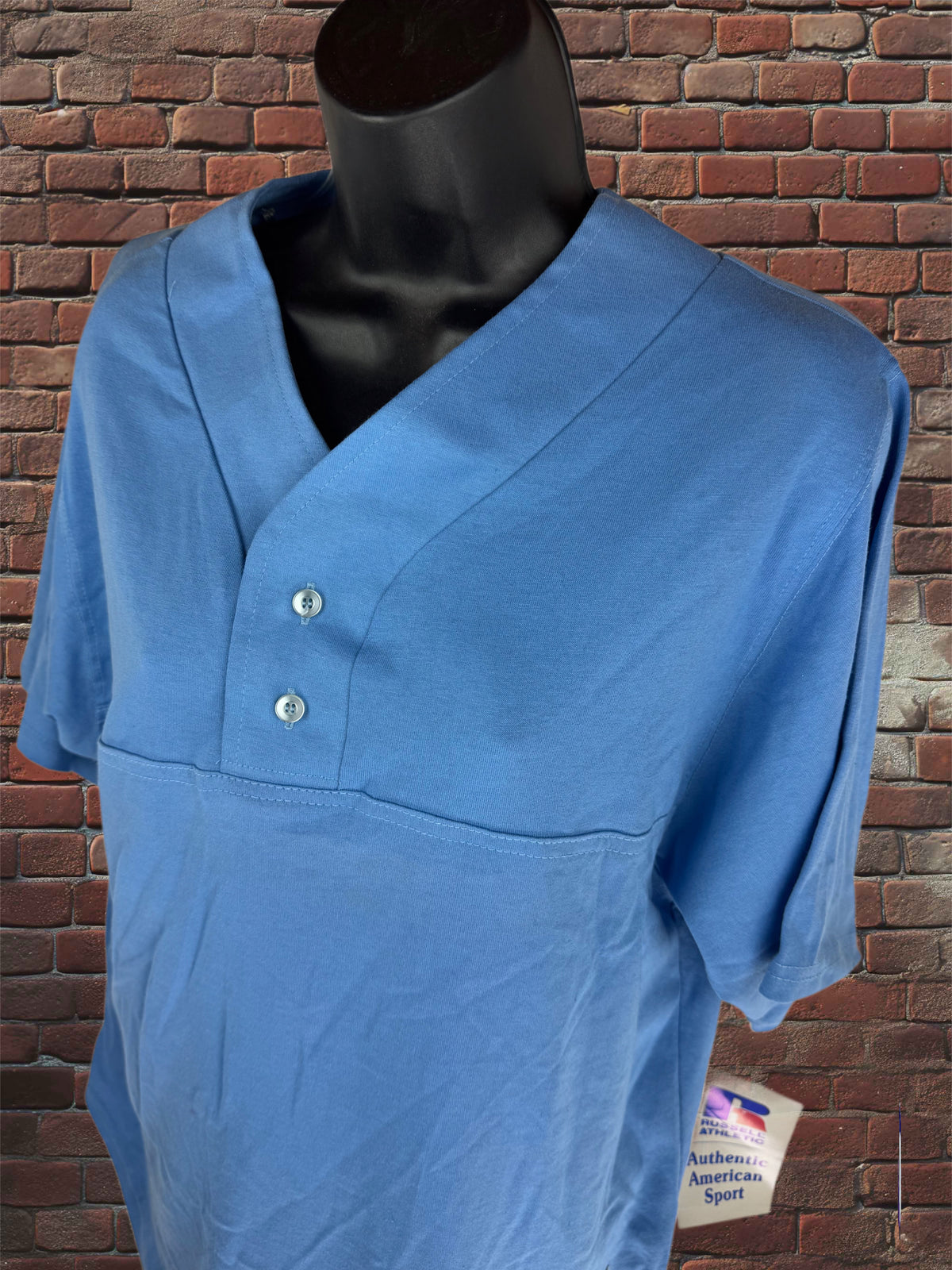 A light blue short-sleeve athletic t-shirt with a split V-neck and two silver buttons, displayed on a black mannequin torso against a red brick wall background.