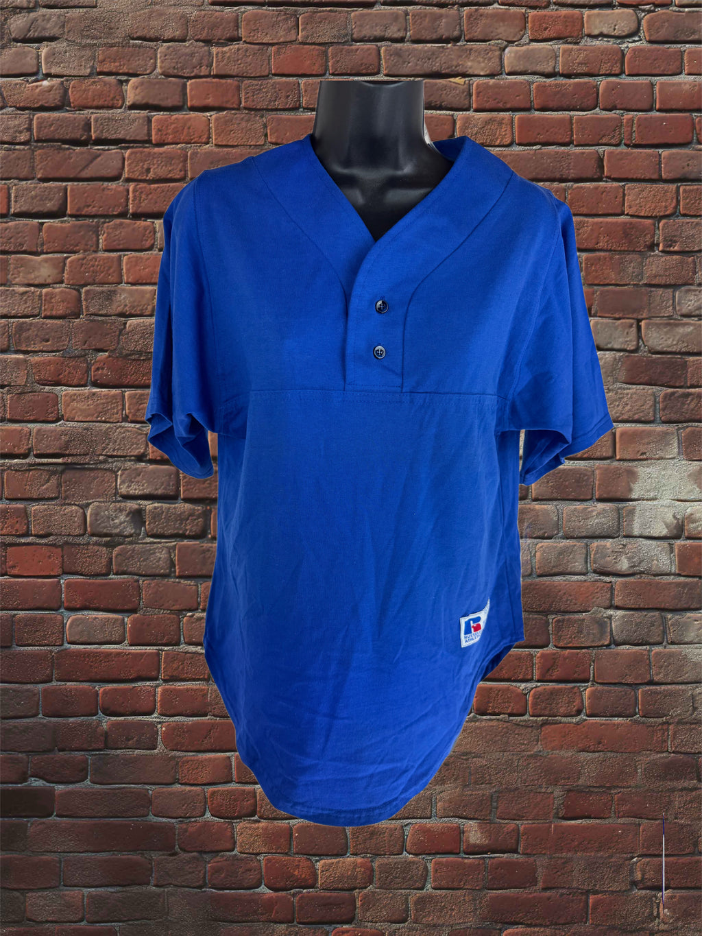 A royal blue short-sleeve baseball-style henley shirt displayed on a black torso mannequin against a red-brown brick wall background.