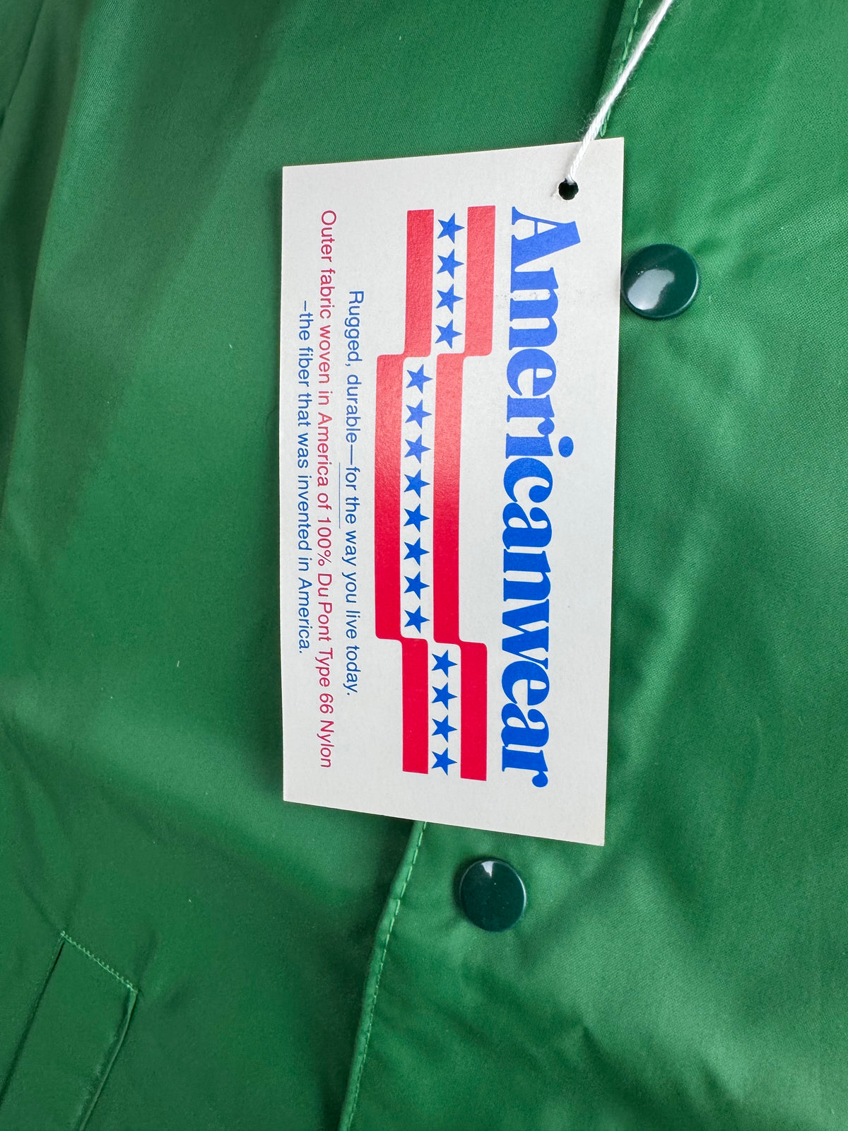 A close-up of a green garment with a white hangtag printed with the brand name 'Americanwear' in blue and red.