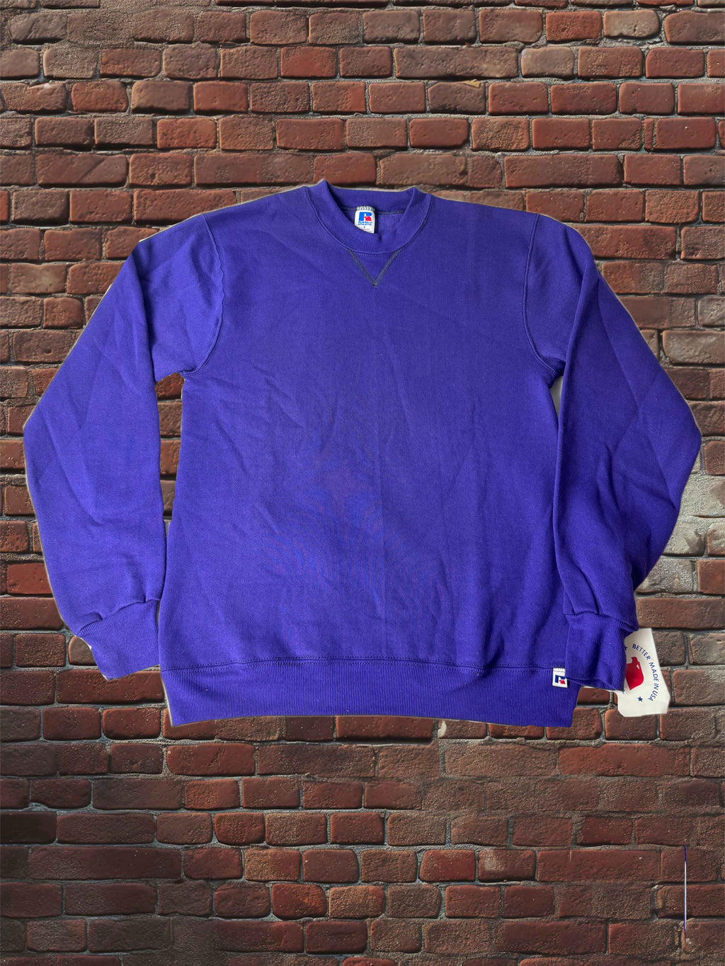 A purple long-sleeve crewneck sweatshirt laid flat against a red brick wall background, with a small white tag attached near the right sleeve cuff.