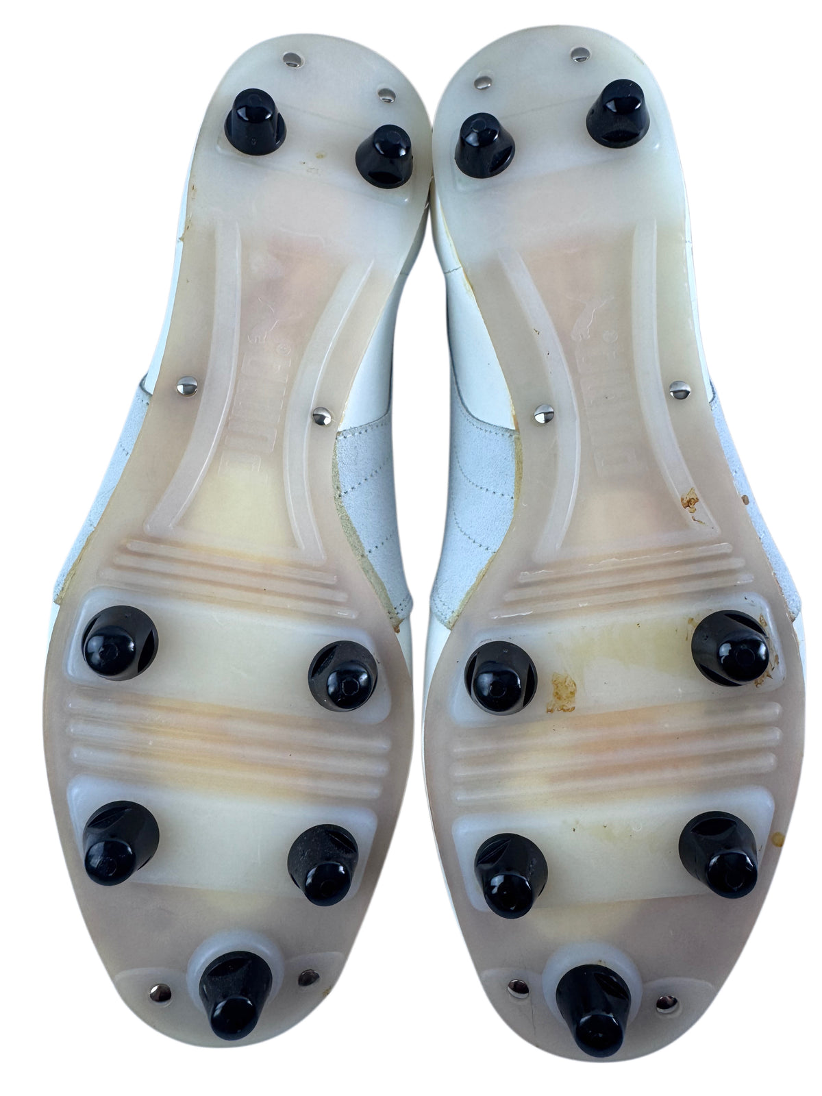 A pair of white athletic cleats shown from the sole, featuring translucent off-white rubber plates with black metal studs, photographed against a solid white background.