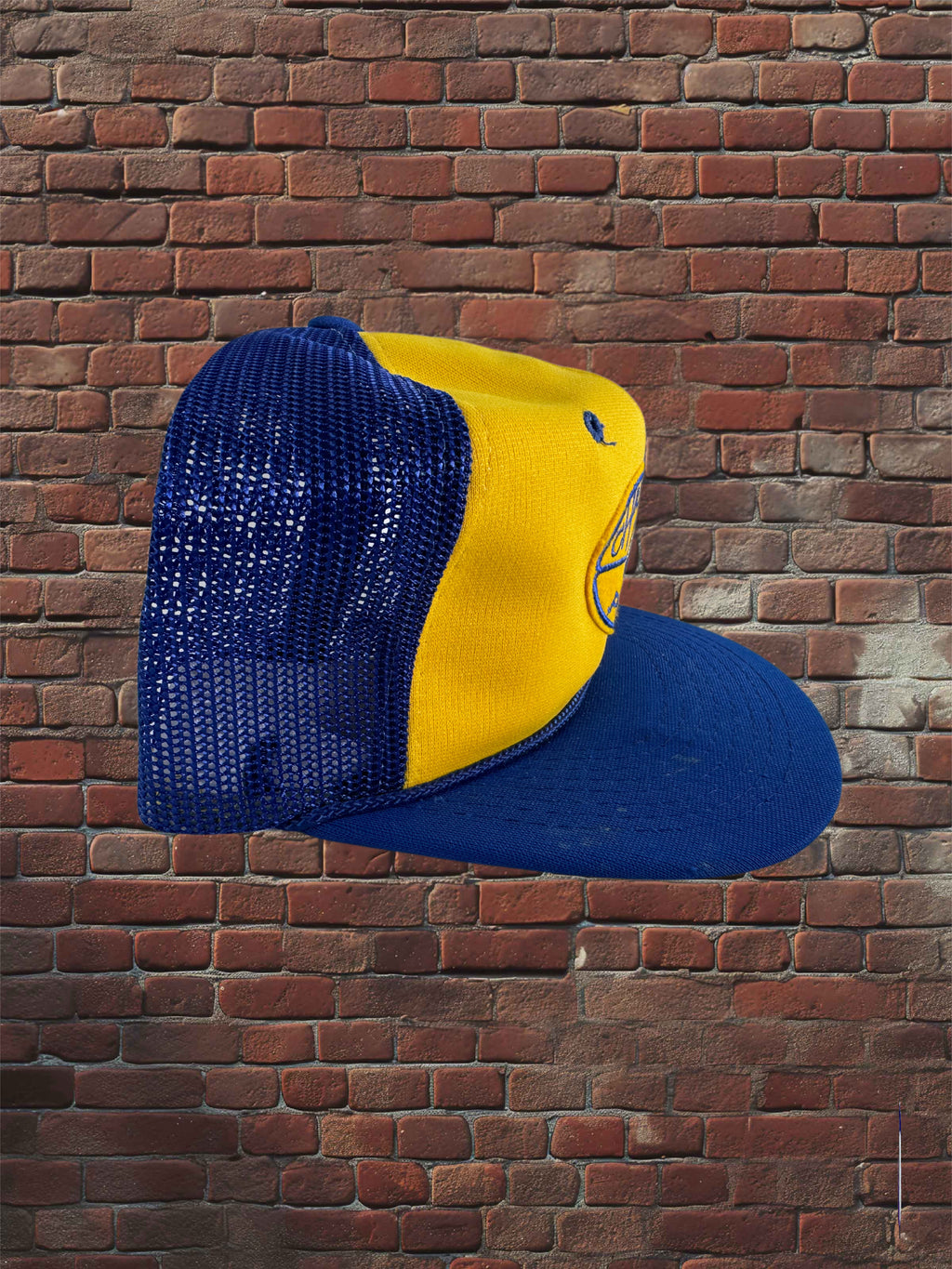 A two-tone trucker cap with a yellow front panel and a blue mesh back and flat brim, shown against a red-brown brick wall background.