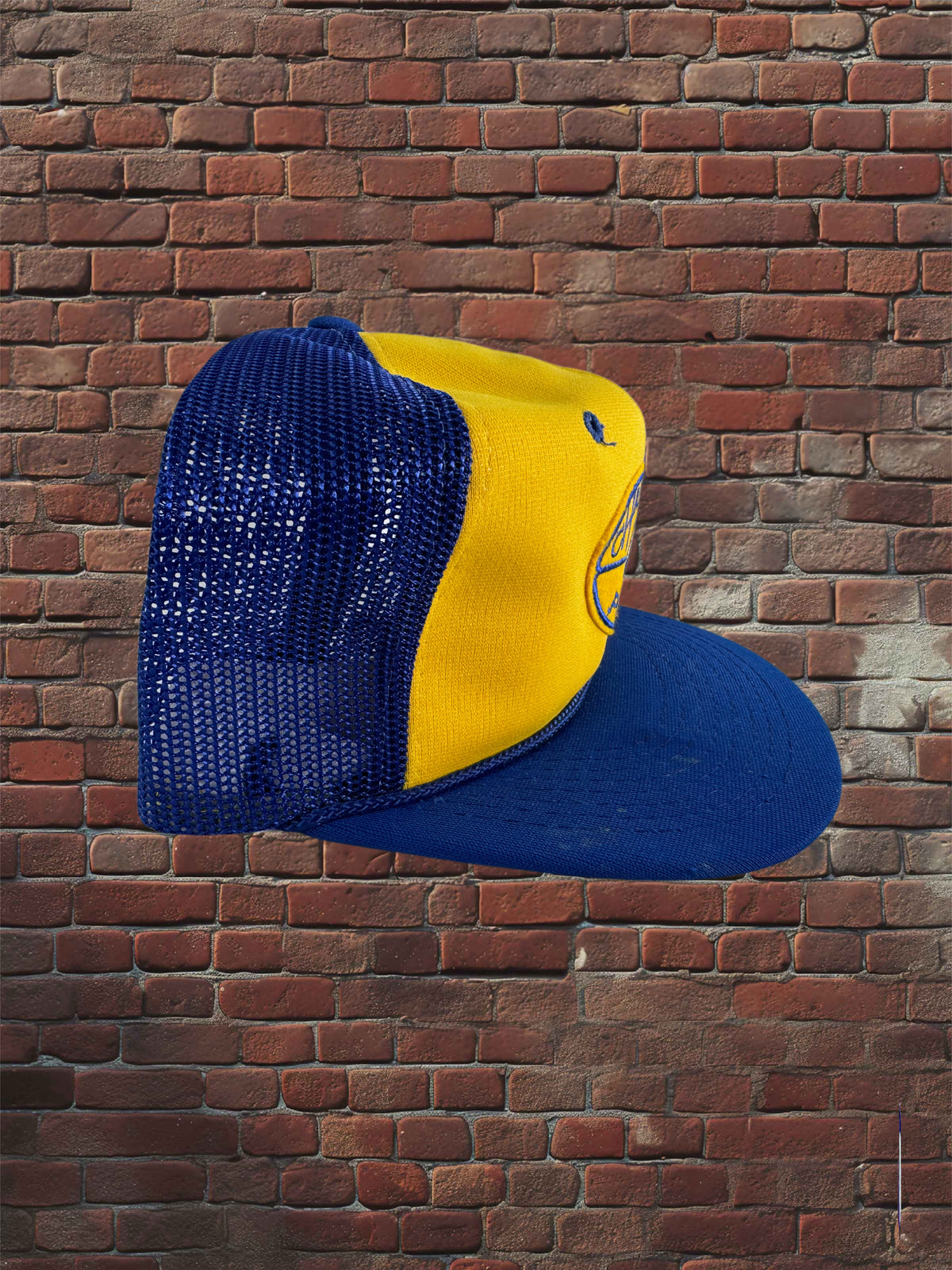 A two-tone trucker cap with a yellow front panel and a blue mesh back and flat brim, shown against a red-brown brick wall background.