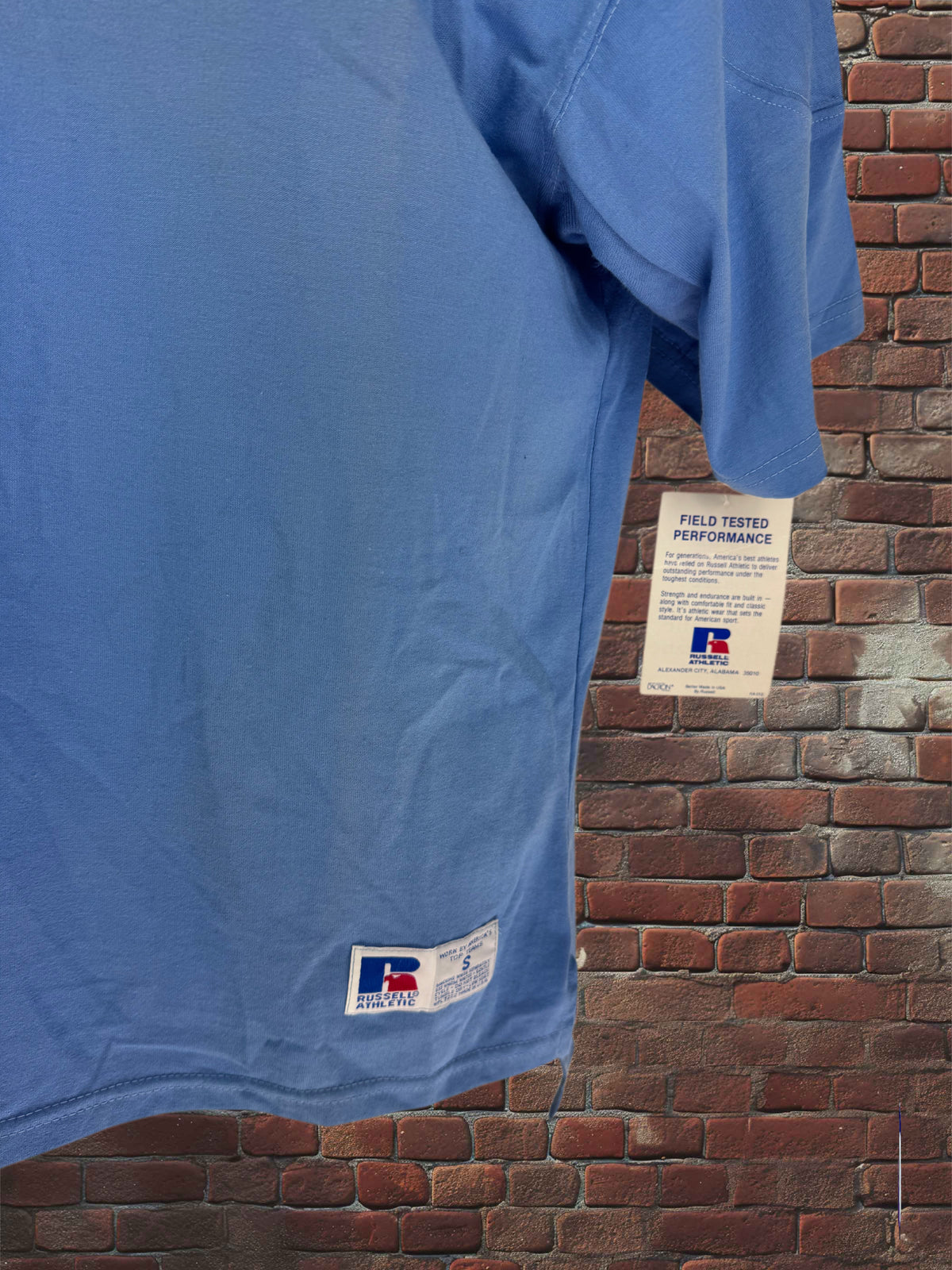 A light blue short-sleeve athletic T-shirt hanging against a red brick wall, with a visible 'Field Tested Performance' tag and a small white brand label near the lower hem.