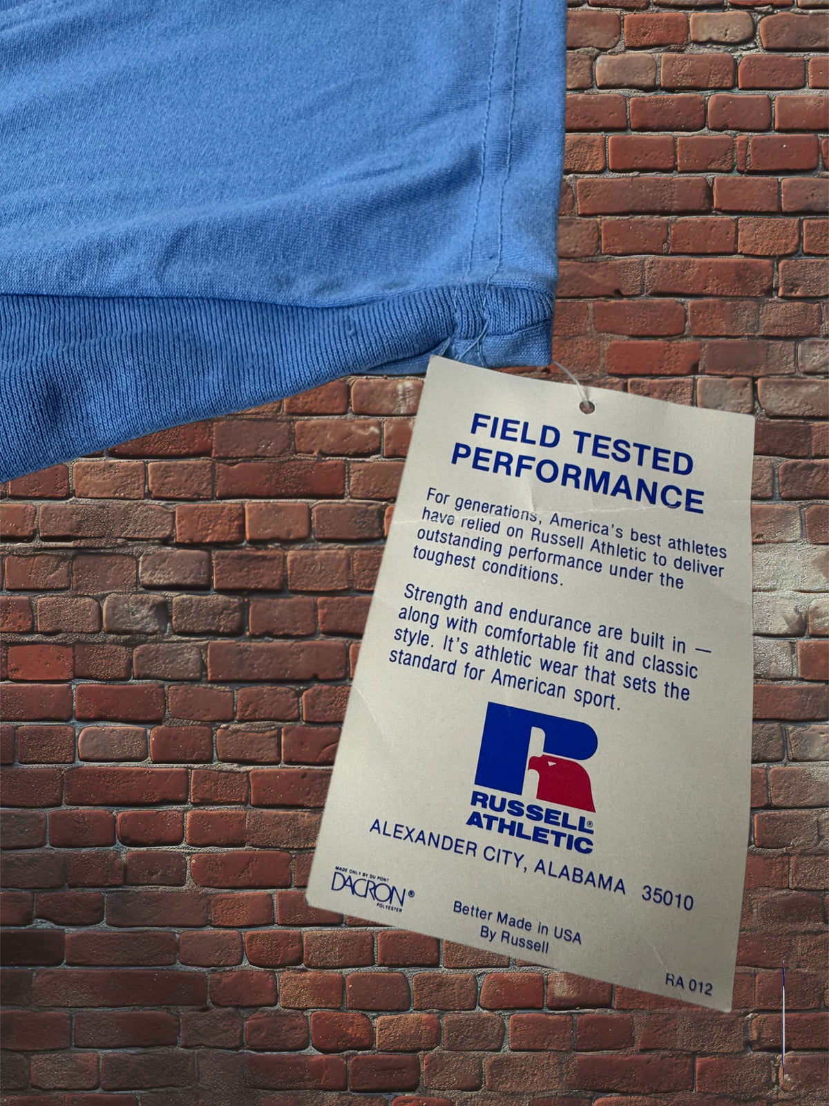 A blue athletic shirt with a white Russell Athletic hangtag, displayed against a red brick wall background.
