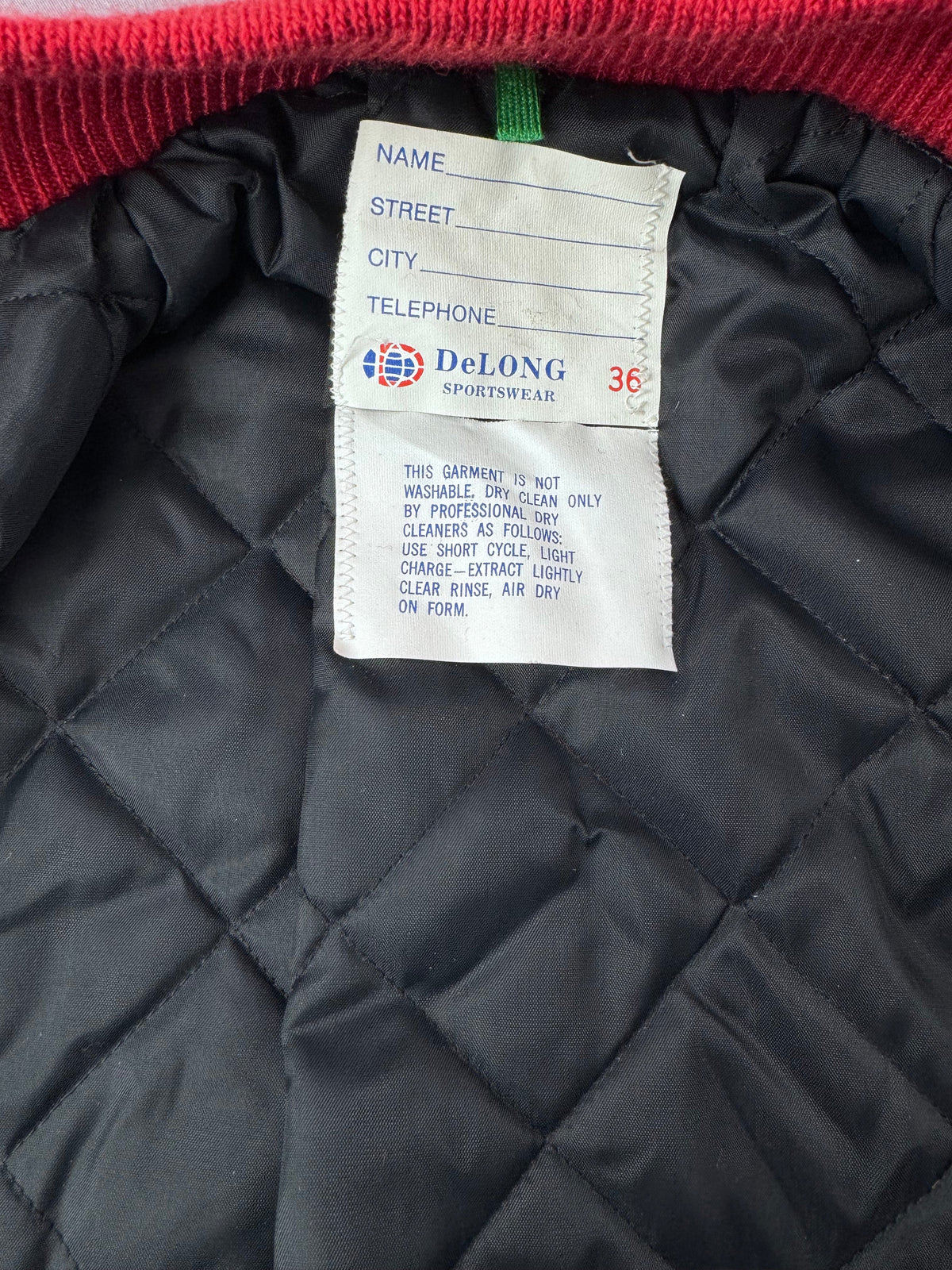 Close-up of a quilted black lettermens jacket interior showing a white DeLong brand sew-in label and a red ribbed knit collar.