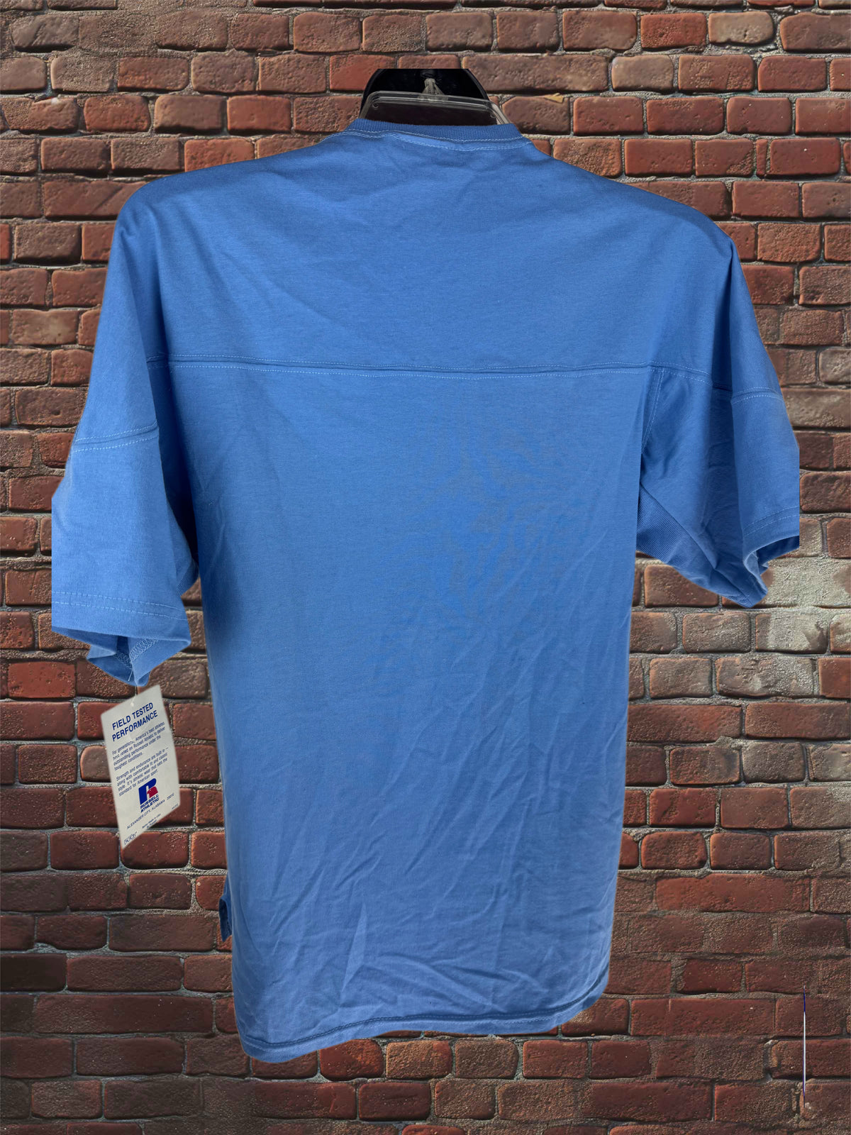 A blue short-sleeve t-shirt hanging on a black hanger against a red brick wall background, showing the back of the garment with a hangtag attached.
