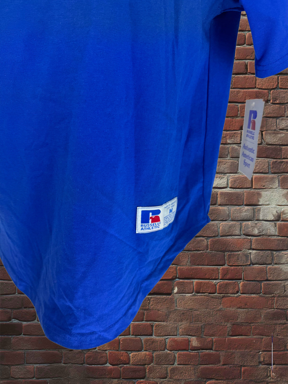 A bright blue short-sleeve shirt hanging against a red brick wall, with a visible white and blue 'R' logo patch near the lower left hem.