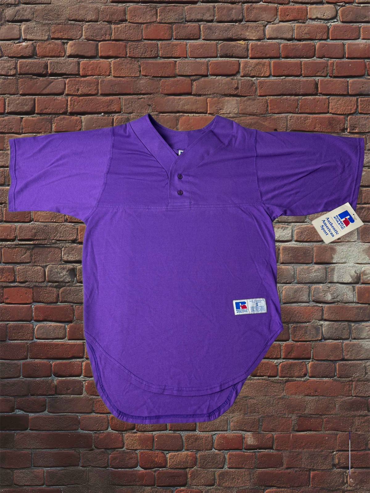 A purple short-sleeve henley-style softball shirt with a curved hem, laid flat against a red-brown brick wall. A small white brand tag is attached near the lower right side.