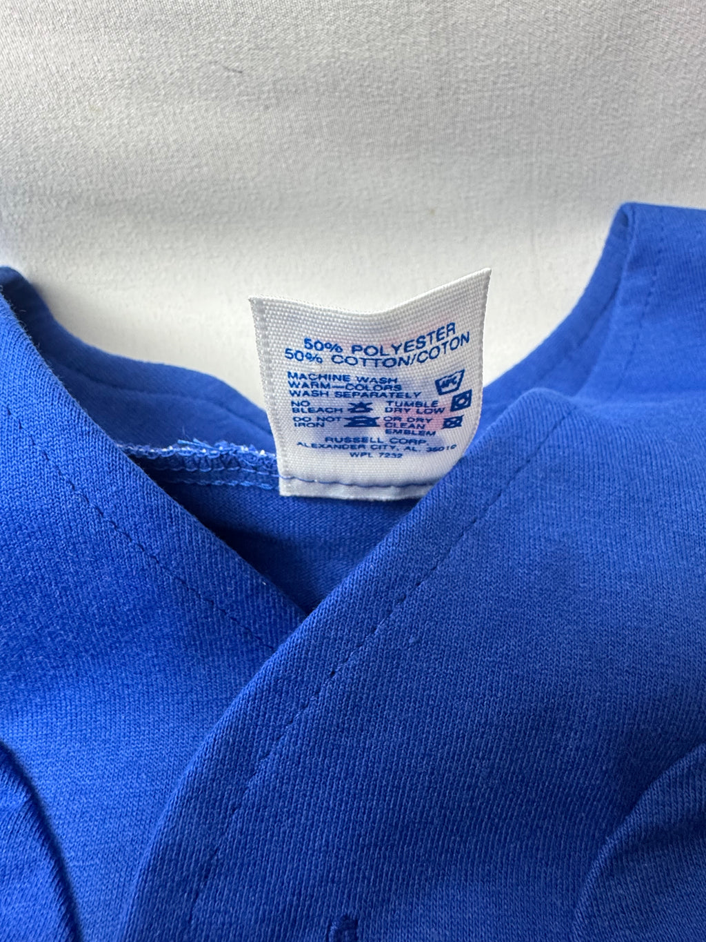 Close-up of a royal blue garment showing the inner neck label, which reads '50% POLYESTER 50% COTTON/COT' with care instructions printed in blue on the white tag.