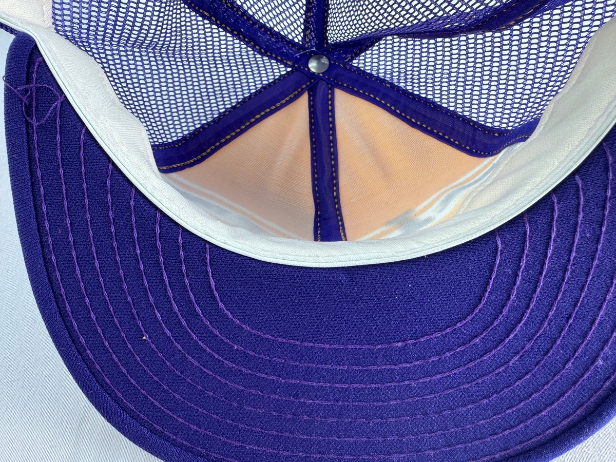 Close-up of the interior back of a trucker-style cap, showing a purple visor, purple mesh back panels, a white fabric band, and peach-colored fabric lining.