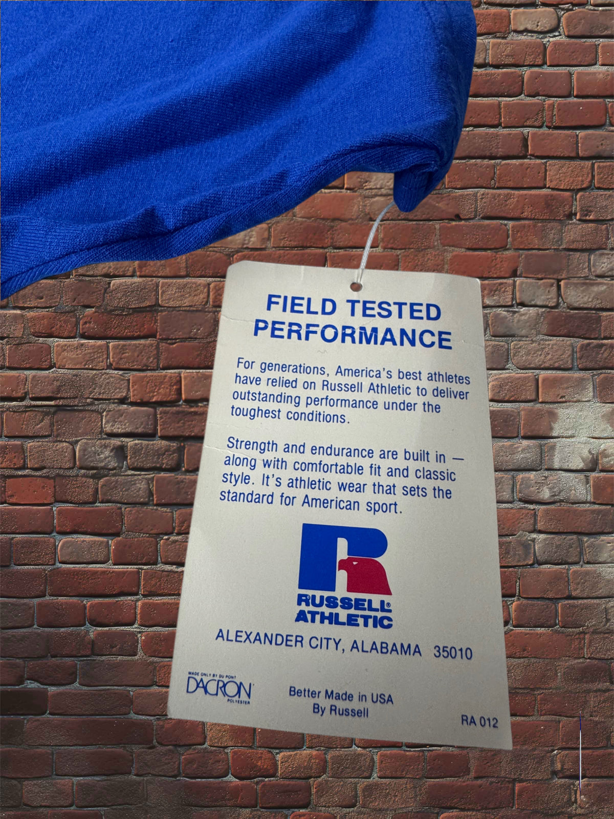 A white paper hangtag with blue text reading 'FIELD TESTED PERFORMANCE' and Russell Athletic branding, attached to a blue garment against a red brick wall background.