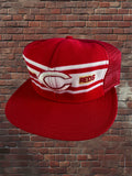 A red and white trucker-style baseball cap with a curved brim, featuring a large embroidered Cincinnati logo and the word 'REDS' on the front panels, set against a brick wall background.