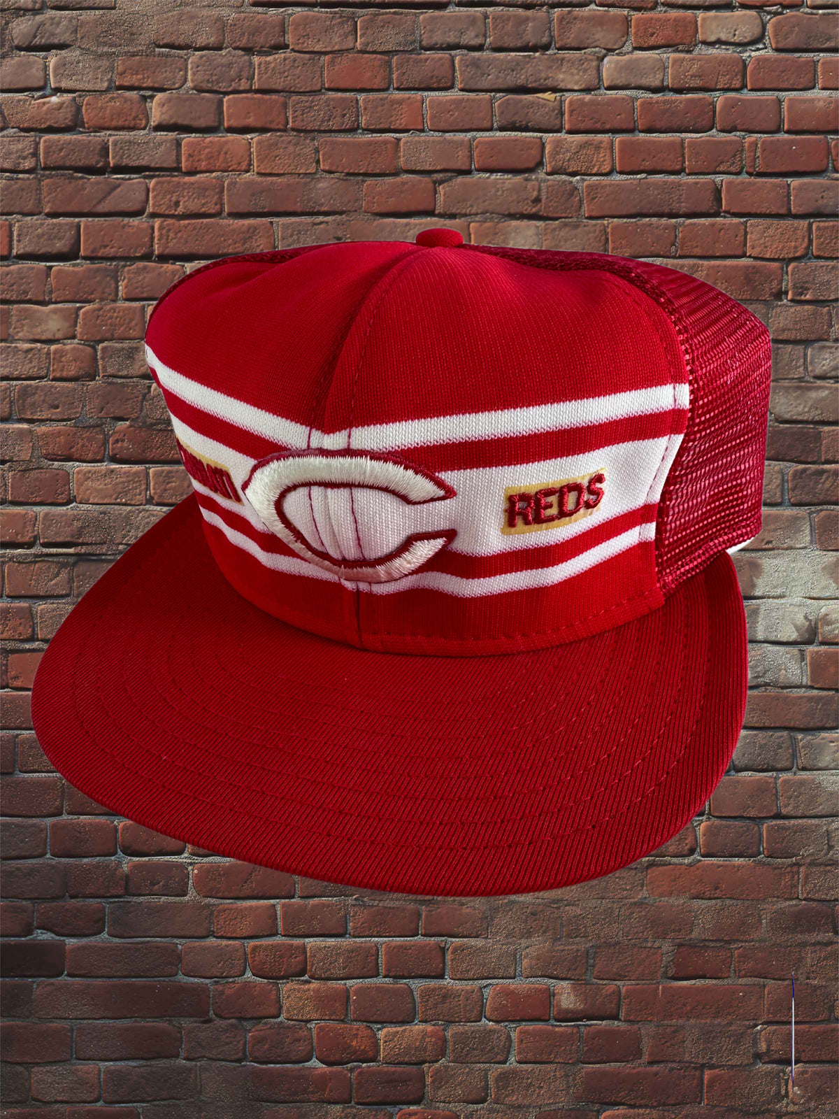 A red and white trucker-style baseball cap with a curved brim, featuring a large embroidered Cincinnati logo and the word 'REDS' on the front panels, set against a brick wall background.