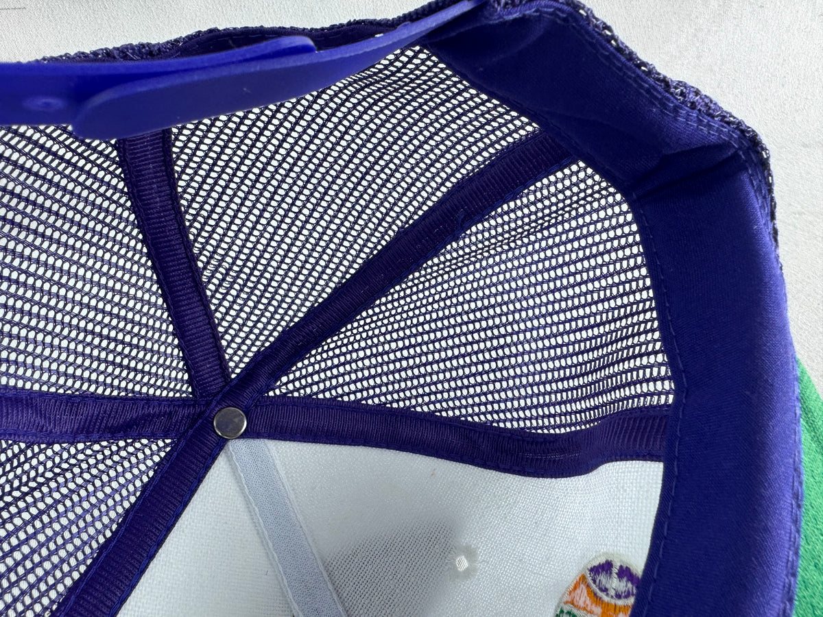 Close-up of the inside crown of a trucker-style cap, showing a purple mesh back, purple fabric brim, and a small multicolor embroidered patch near the lower right edge.