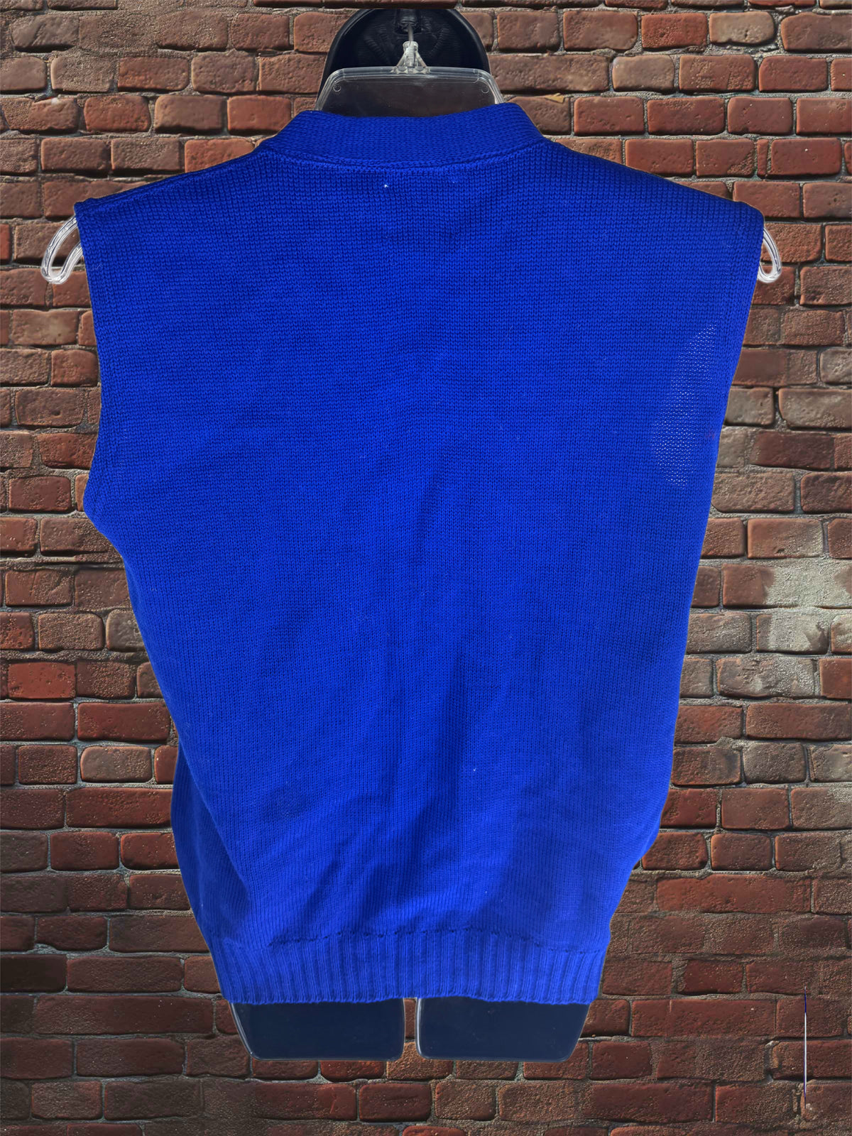 A bright royal blue sleeveless knit sweater vest displayed on a black hanger against a red-brown brick wall background.