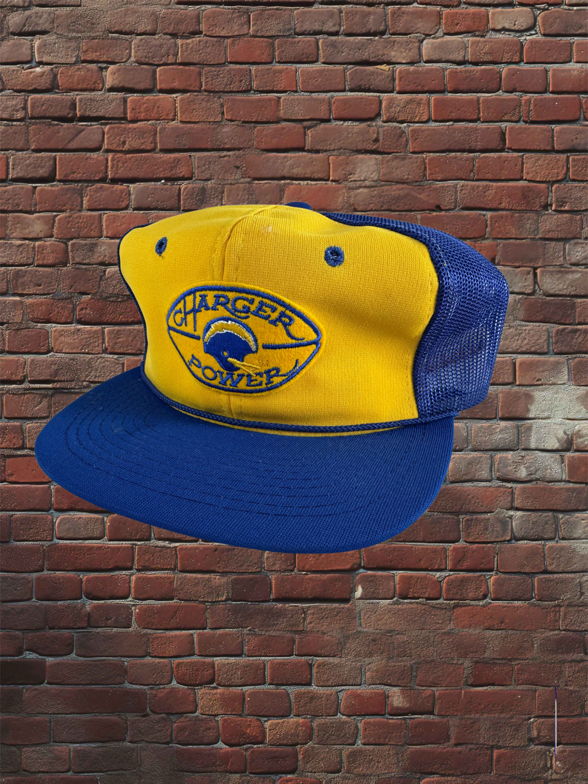 A yellow and blue trucker-style cap with a curved blue brim, shown against a red-brown brick wall. The front panel is yellow with a blue and white 'Charger Power' logo, and the back mesh is blue.