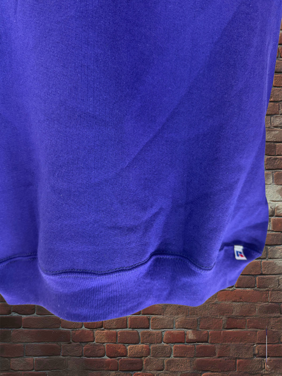 A close-up of a vibrant purple sweatshirt hanging against a rustic red brick wall background. The fabric is shown with soft folds and a ribbed hem, featuring a tiny white and blue tag on the lower right side.